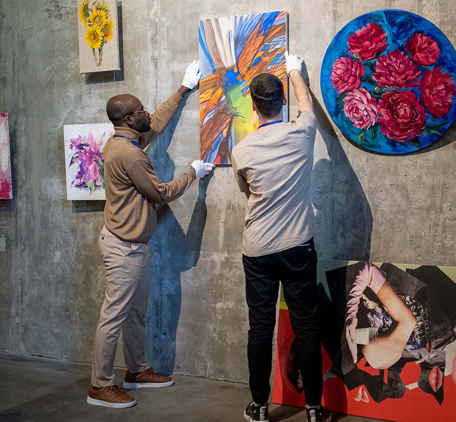Two individuals carefully hanging colorful artwork on a concrete wall in a gallery.