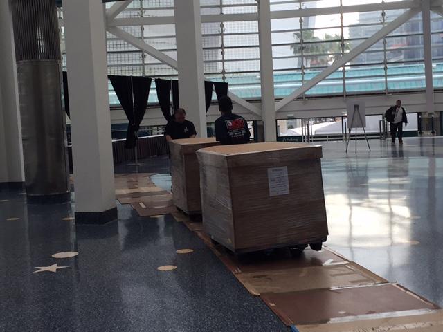 Two workers moving large wooden crates on a cart in a spacious venue.